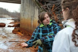 Woman (Gabby Zawacki) displays a rust-colored sludge on her fingertips to unseen group of adults. She is in front of concrete culvert over river, which is also orange.
