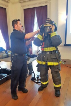 Man in navy work uniform assists woman in trying on firefighter suit and gas mask