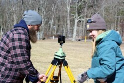 A man on left and woman on right work to level a tripod with target on it in field with leafless trees behind them
