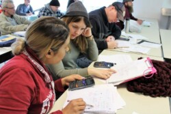 Two women at closest edge of long desk work on calculator with notebooks and worksheets in front of them. Other students next to them and in other rows also work on calculators