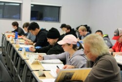 Adults, seated at two long rows of desk, look at binders. Most hold pens.