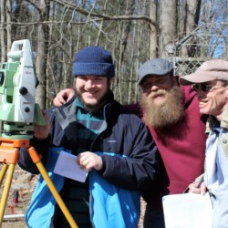 One man in center puts arms around shoulder of two other men; one the left side is a piece of surveying equipment (total station)