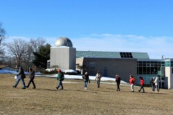 Line of 11 adults walk across field with patches of snow. A concrete building with large windows and a small observatory dome are in background.