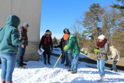 Several adults in winter jackets look down into snow. One person holds a yellow stick (metal detector).