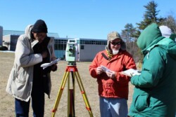 Four men in winter coats stand around total station on tripod outside on field on college campus