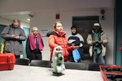 Instructor in heavy winter coat talks while holding a piece of equipment (total station) on table. Four adult students, also in coats, listen to her.