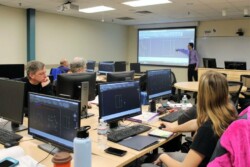 Instructor at front of classroom points to projection of his computer screen with rectangular schematic on it while four students seated at computers look on