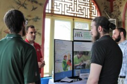 Man stands to right of a tabletop display and speaks to three men facing him