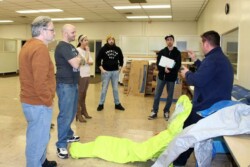 Five adults standing on left listen to instructor on right, who is holding a fluorescent yellow hazmat suit
