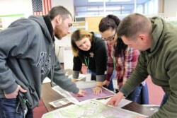 Male adult stands on one side of table pointing down to location on large map; three other adults (two women, one man) stand on opposite side, also looking at or pointing to map.