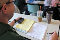Looking down over shoulder of man, with pen in hand, seated at long desk. Several notebooks with geometric calculations are in front of him, as well as two coffee drinks.