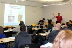 Classroom with adult students sitting at long tables looking at Powerpoint slideshow, while a male instructor, at right, gestures towards screen