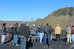 Landscape view of one man in front of a large pile of mining waste, talking to group of adult students (looking at bank).
