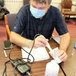 White male adult with notebook sitting at table, inserting a probe into a coffee cup. Probe is attached to cord and electronic/digital device.