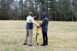Man (Frank Derby) on left speaks to adult student on right with a yellow surveying tripod in between them