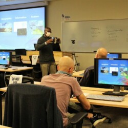 Instructor in front of room, talking and gesturing with hands, speaks to adult students at computers, with screens reading "Geographic Information System"