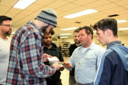 One adult male flips through small, spiral-bound book, with five other men look on or discuss it with him