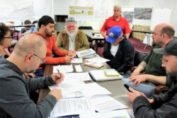 Seven adults site around table covered with books, papers, and notebooks working, while a seated male instructor looks on