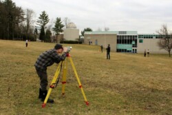 Man peering through level on tripod on grassy field with block concrete college building with large windows in background