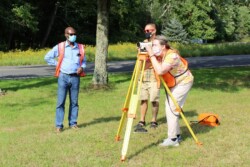 Woman peers through level on tripod while two men to her right side look on outdoors on green grass