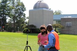Man and woman looking at clipboard next to tripod with yellow and black target on top, outdoors on a college campus